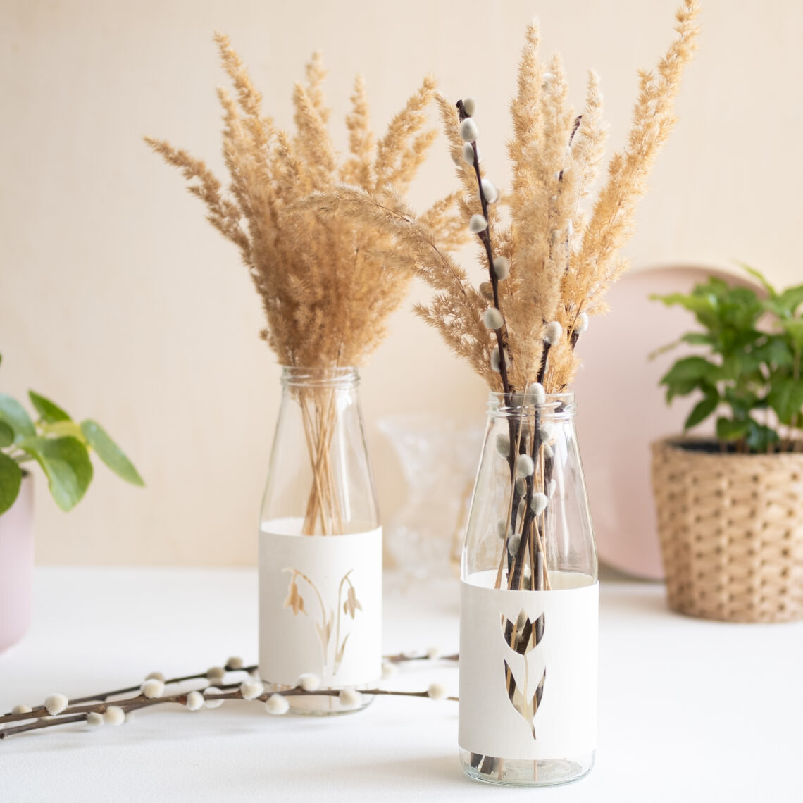 Zwei DIY upcycling Frühlingsvasen aus Altglas sind mit Zweigen und trockenen Gräsern befüllt und dienen als Tischdeko