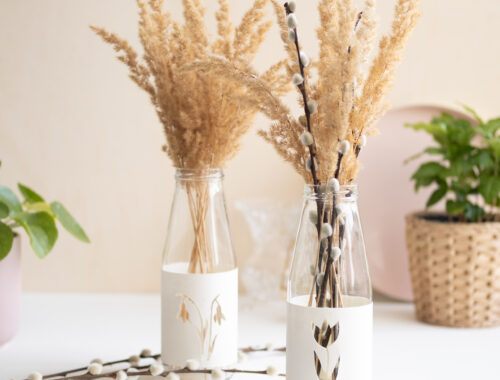 Zwei DIY upcycling Frühlingsvasen aus Altglas sind mit Zweigen und trockenen Gräsern befüllt und dienen als Tischdeko